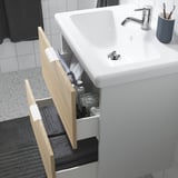 Lavabo de bany blanc amb calaix de roure obert amb articles de tocador. Aixeta cromada i suport de raspall de dents gris a la dreta.