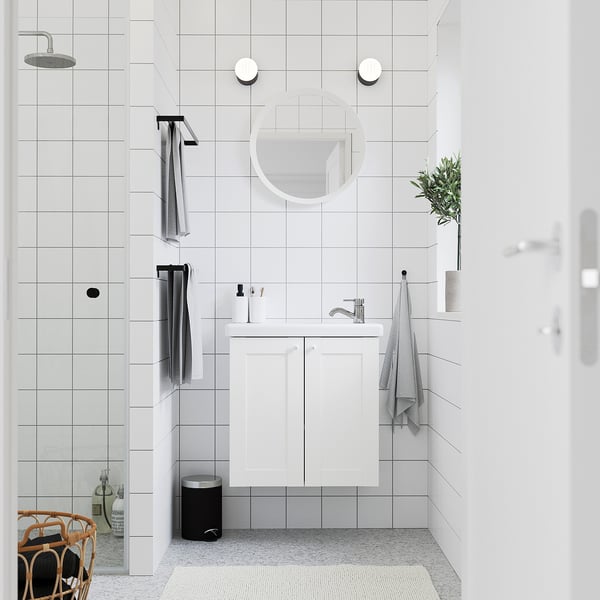 Bany modern amb parets blanques i accessoris. Armari ENHET blanc, mirall rodó, dutxa. Tovallola grisa, planta petita. Brillant, minimalista.