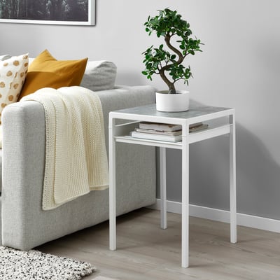 Petita taula auxiliar blanca amb una planta artificial compacta FEJKA a la part superior, al costat d'un sofà gris.