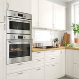 Una cuina moderna amb forn encastat d'acer inoxidable FORNEBY i microones. Els armaris blancs l'envolten, amb un taulell de fusta i un respatller de rajoles.