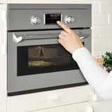 Persona que utilitza un modern forn doble d'acer inoxidable amb pantalla digital i controls tàctils en una cuina blanca.