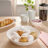 Un bol d'ous de colors barrejats es troba a la taula del menjador envoltat de pa, tasses de cafè i fruita, que suggereix l'esmorzar.