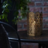 Una llanterna decorativa s'asseu sobre una taula a l'aire lliure, emetent una brillantor càlida a través del seu intricat patró floral. El mànec llarg permet un fàcil transport.