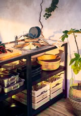 Lavabo exterior GRILLSKÄR negre amb prestatges d'acer, cistelles de vímet i parament de cuina, col·locat contra una paret lleugera amb plantes.