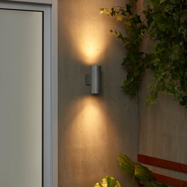Aplic d'exterior gris modern GRÖNSPRÖT sobre paret beix prop de la porta blanca, amb brillantor càlida i contrast de verd.