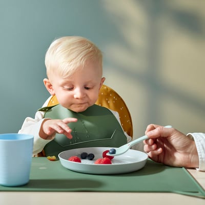 Un nen petit amb un pitet de silicona verd amb una butxaca groga, menjant baies d'un plat blanc amb un adult alimentant-los amb una cullera blava clara.