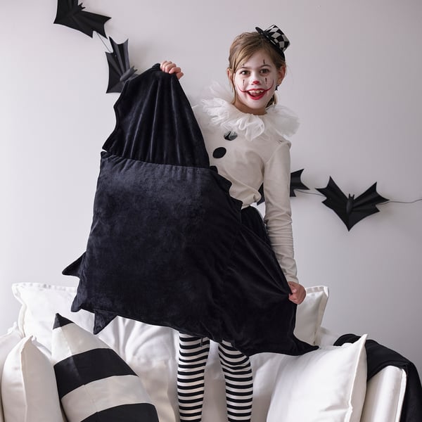 Nen amb disfressa de ratpenat de Halloween sostenint un coixí suau negre, assegut en un sofà blanc amb coixins de ratlles blanques i negres.