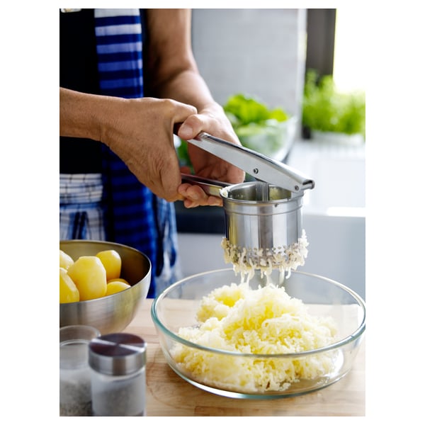 Persona que utilitza premsa de patates d'acer inoxidable per triturar patates bullides en un bol. Taulell de cuina amb llimones, sal i herbes visibles.