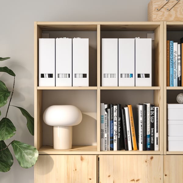 Prestatge d'emmagatzematge de fusta amb compartiments ajustables, amb llibres i arxius. Una làmpada es troba en un prestatge del mig. Es col·loca una planta al costat de la prestatgeria.