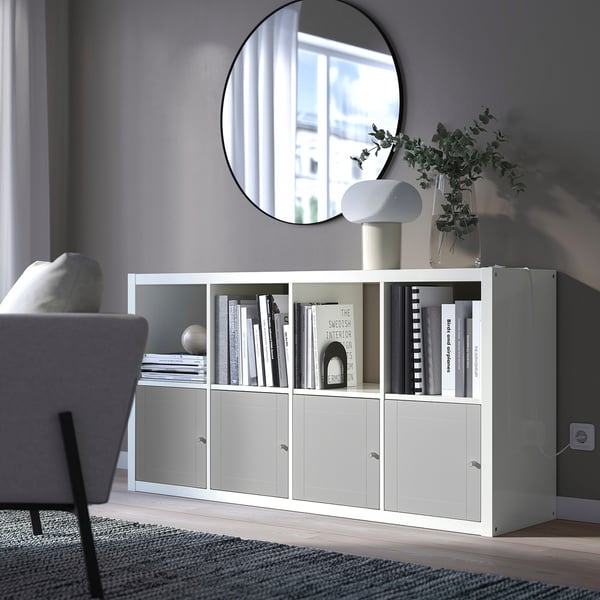 Prestatgeria KALLAX blanca minimalista, gran, amb llibres i decoració, s'adapta sota un mirall rodó, al costat d'una cadira acollidora.