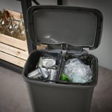 Contenidor de pedal negre amb dos compartiments interiors, un amb llaunes i l'altre amb residus plàstics, dissenyat per a la separació de residus.