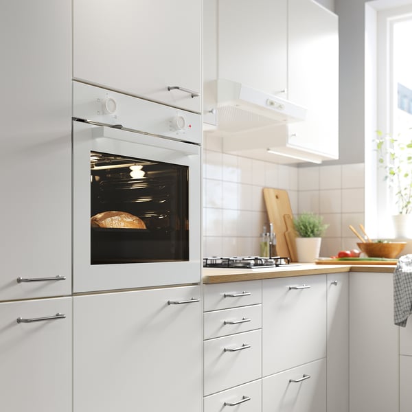 Cuina blanca moderna amb forn incorporat, cuina de gas, taula de tallar de fusta i productes frescos al taulell.