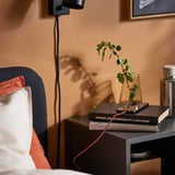 Configuració de nit amb un cable USB LILLHULT negre que carrega un telèfon a sobre de llibres, a prop d'una planta i un aplic.
