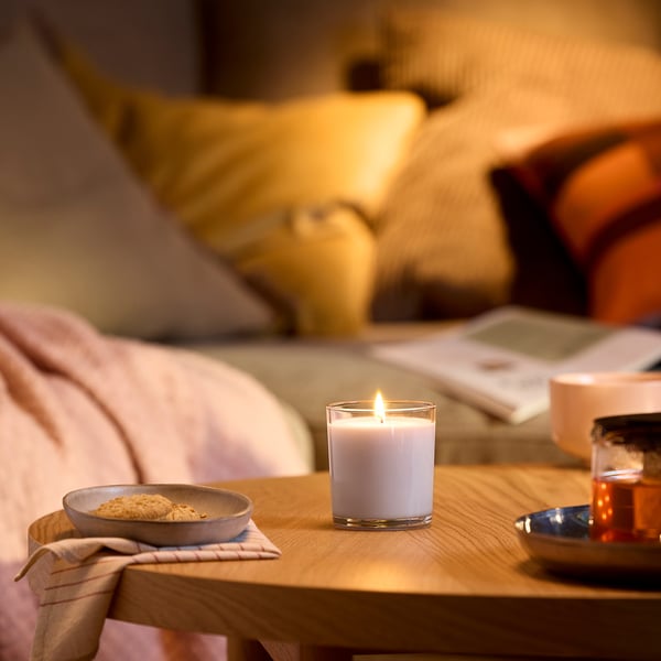 Habitació acollidora amb espelma blanca LOTSFÅGEL sobre safata de fusta. Bol, drap, llibre i got a prop. Ambient tranquil, il·luminació suau.