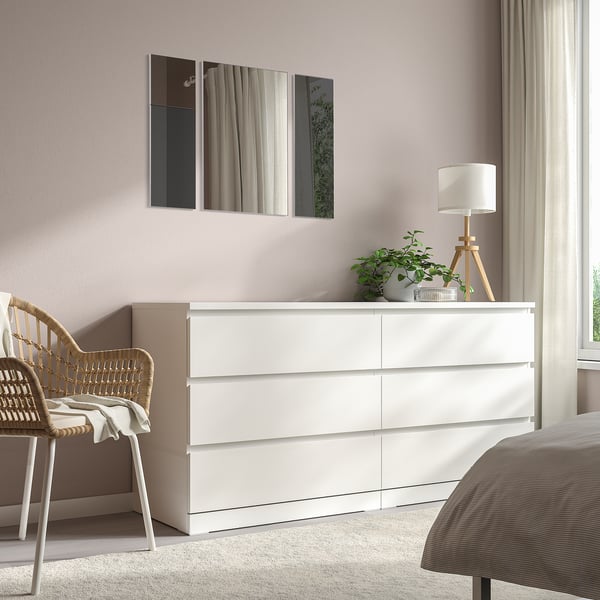 Un dormitori modern amb aparador blanc MADESJÖ, alt i rectangular, rematat amb llum de taula i planta. Mirall i cadira de vímet al costat.