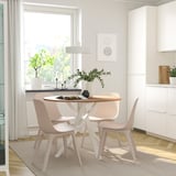 Un menjador modern amb taula rodona MARIEDAMM i cadires de color rosa clar, envoltat d'un gran finestral, testos de plantes i una decoració minimalista.