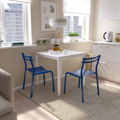 Conjunt de menjador modern i lluminós amb taula blanca i cadires blaves contra un gran finestral. Compacte però elegant, que s'adapta perfectament a la sala petita i lluminosa.