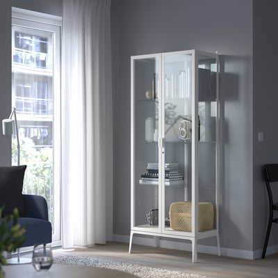 Armari de portes de vidre blanc amb llibres i decoració, prop de la cadira blava, habitació tranquil·la.
