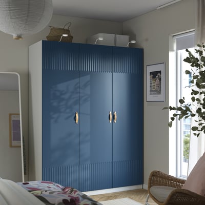 Dormitori modern amb armari PAX blau, cistelles d'emmagatzematge, cadira de vímet, mirall, llit floral i planta penjant.