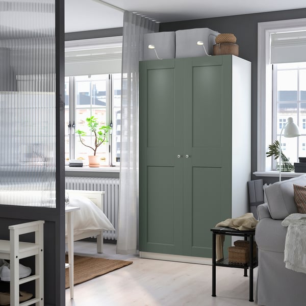 Aquesta imatge és un armari PAX verd en una habitació moderna. Es troba alt, al costat d'un sofà i una prestatgeria, oferint un ampli espai d'emmagatzematge amb les seves dues portes.