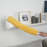 La mà sosté el plomall de microfibra groc, netejant sobre el sofà. Llum de paret blanca i prestatgeria amb art emmarcat al darrere.