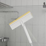 Un mànec de neteja telescòpic blanc amb un racó groc i blanc, netejant una paret de dutxa amb gotes d'aigua.