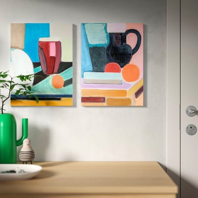 Dos llenços abstractes de colors pengen sobre una taula de fusta amb un gerro verd, plats blancs i una petita jardinera.
