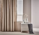 Sala d'estar minimalista amb cortines beix, consola grisa, gerro blanc amb branques i manta drapejada.