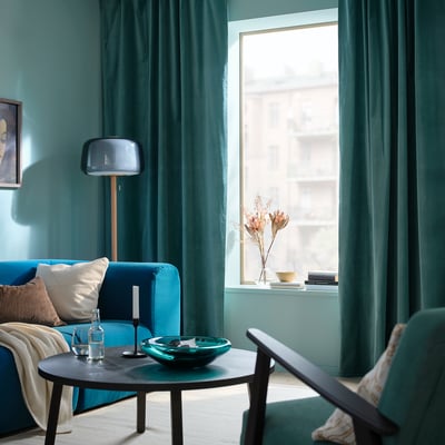 Dues cortines SANELA de color gris turquesa fosc pengen una al costat de l'altra, amb una luxosa textura de vellut i un disseny de capçalera multidireccional.