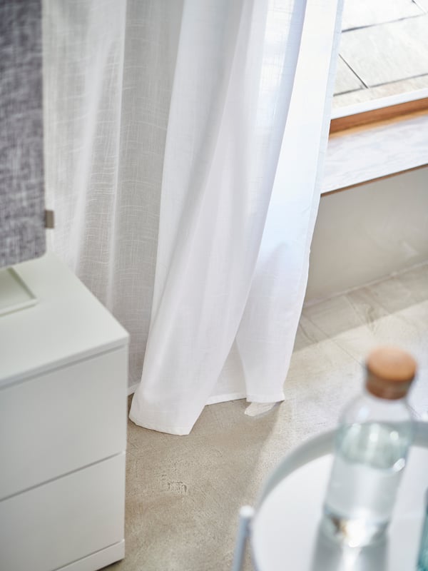Les cortines blanques pengen al costat d'un armari blanc. La llum es filtra suaument a través de la textura semblant al lli del teixit 100% cotó, creant ombres suaus.