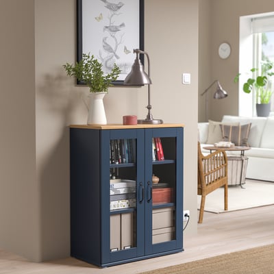 Armari SKRUVBY blau amb portes de vidre, tiradors rústics i planta a prop.