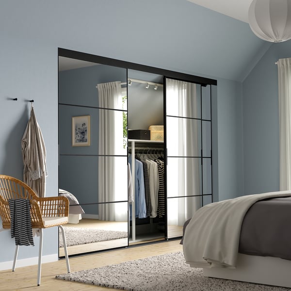 Dormitori blau cel modern amb portes d'armari miralls SKYTTA que milloren l'amplitud, marcs negres, roba de llit blanca, cadira de vímet i decoració neutra.