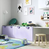 Habitació infantil amb caixa de joguines SMÅSTAD, morat clar, rectangular, guarda les joguines ordenadament. És més gran que el tamboret del costat, s'adapta a l'art de la paret penjant.