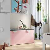 Habitació infantil amb banc d'emmagatzematge de joguines SMÅSTAD blanc de color rosa clar, amb joguines de dinosaures i estampats artístics de colors a la paret.