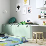 Habitació infantil amb banc verd SMÅSTAD que emmagatzema joguines, catifa de colors, tamboret de fusta i art mural. Disseny brillant i lúdic, perfecte per jugar.