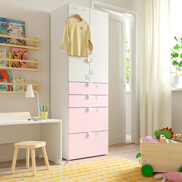 Una habitació infantil amb un armari SMÅSTAD blanc i rosa, prestatges de fusta amb llibres de colors, un petit escriptori i una catifa groga.