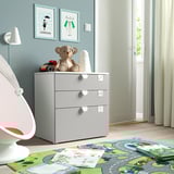 Habitació infantil moderna amb parets de color blau clar. Cofre blanc amb 3 calaixos, rematat amb decoració d'ós de peluix i mussol. La catifa del sòl té un disseny de full de ruta.
