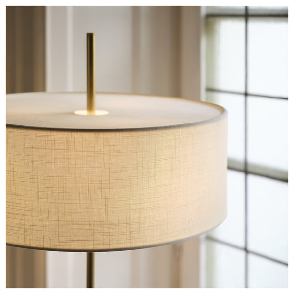Primer pla d'un elegant llum de peu amb una pantalla beix i textura i un suport de llautó, que reflecteix materials d'alta qualitat i un disseny elegant.