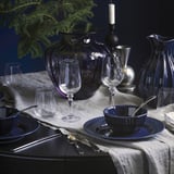 Elegant configuració de sopar amb cristalleria blava i clara, gerros i estris de menjador disposats sobre estovalles blanques. Les espelmes i la vegetació decoren la taula.