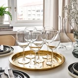 Sis copes de vi clar en una safata daurada al costat de plats i coberts, col·locades prop d'una finestra amb cortines blanques.