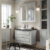 Bany gris modern amb tocador TÄNNFORSEN, mirall gran i lavabo elegant. Plantes i tovalloles decoren l'espai.