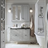 Bany gris modern amb armaris de fusta clara, banyera blanca, lavabo i mirall. Els armaris de paret estalvien espai, terra de rajoles blanques. Il·luminació suau.
