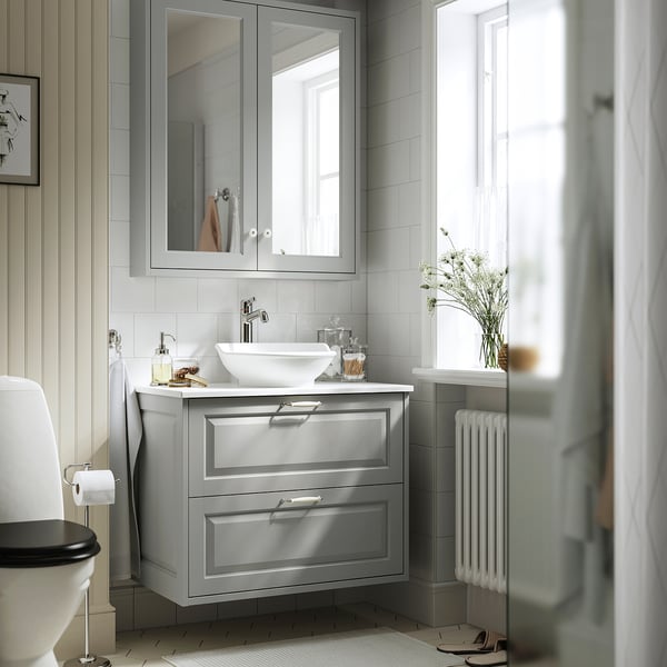 Bany gris modern amb tocador TÄNNFORSEN, lavabo blanc, armari amb mirall i seient de vàter fosc. Tovalloles blanques, flors, tacte net.