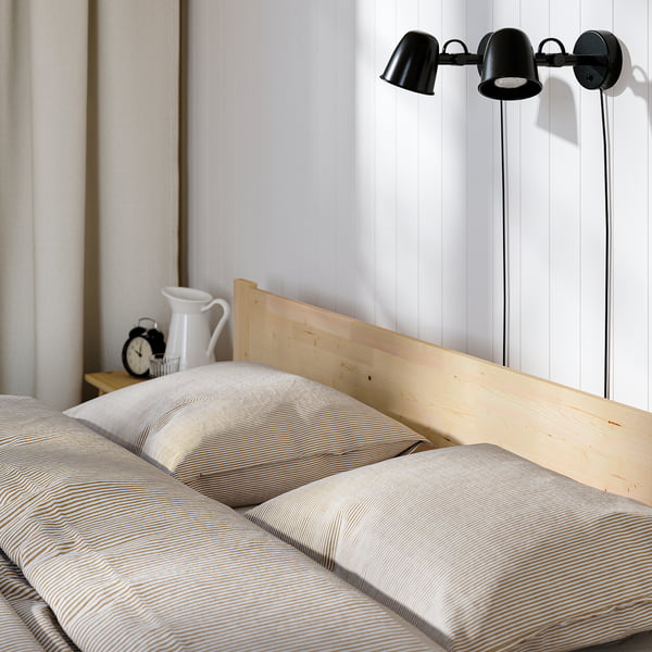 Escena de dormitori amb un marc de llit de fusta, dues làmpades negres muntades a la paret, un rellotge i una gerra blanca sobre una tauleta de nit.