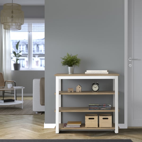 Sala d'estar moderna amb consola TRÄKUMLA, paret grisa, terra de fusta. Planta petita, llibres, rellotges i caixes a les prestatgeries. Porta blanca al costat.