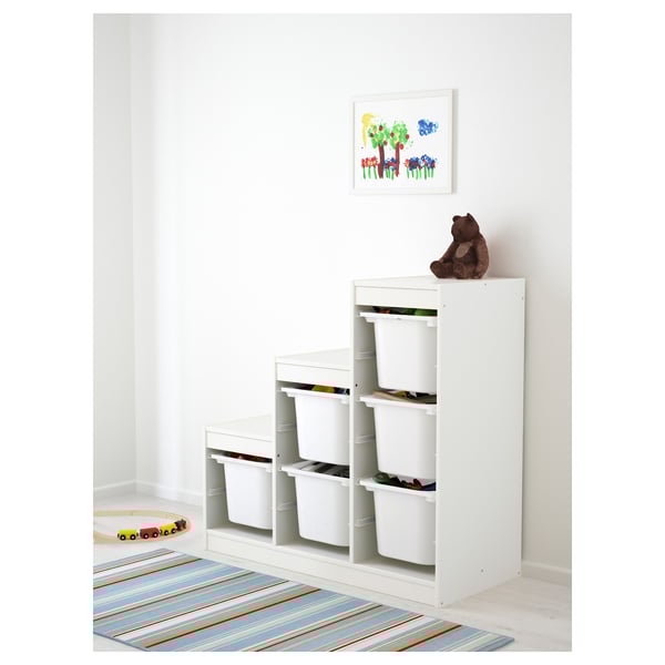 Moble d'emmagatzematge TROFAST blanc amb múltiples contenidors, perfecte per organitzar joguines infantils. Es col·loca un ós de peluix a la part superior i a sota hi ha una catifa de colors.
