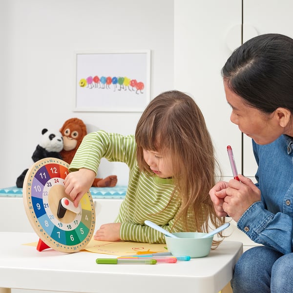 El nen i l'adult s'asseuen a taula, utilitzant un rellotge educatiu i pintar. Les agulles dels rellotges es mouen, mostrant el temps d'aprenentatge.