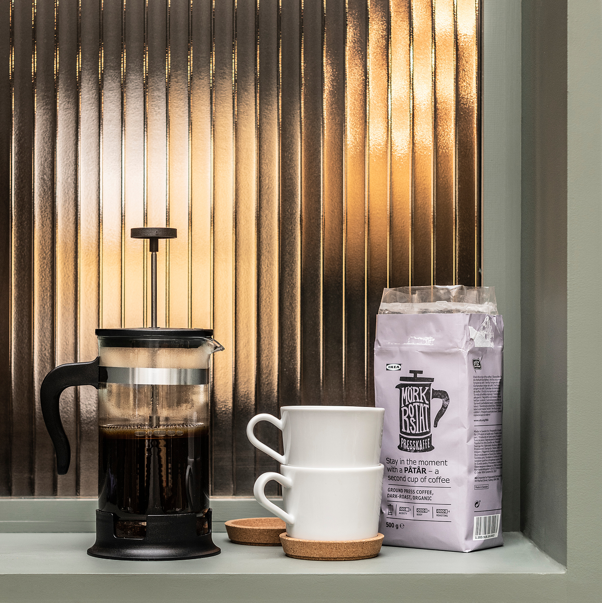 Cafetera de premsa francesa negra amb tasses blanques i bossa de cafè a l'ampit de la finestra.