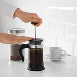 La persona pressiona l'èmbol de la cafetera negra sobre el taulell blanc; tassa i bidó a prop.