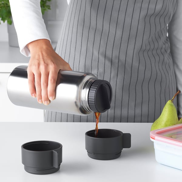 Persona abocant cafè d'un termo metàl·lic a una tassa negra sobre una taula blanca, amb una altra tassa i un recipient de menjar a prop.